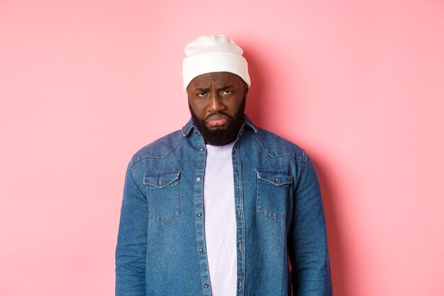 Disappointed and sad Black man, sulking and whining, looking at camera with offended grimace, standing over pink background