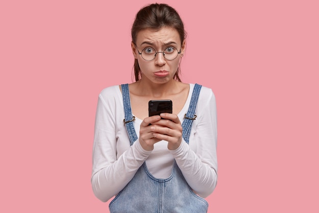 Disappointed sad female purses lower lip in displeasure, doesnt recieve call from boyfriend after quarrel, holds mobile phone, looks desperately at camera