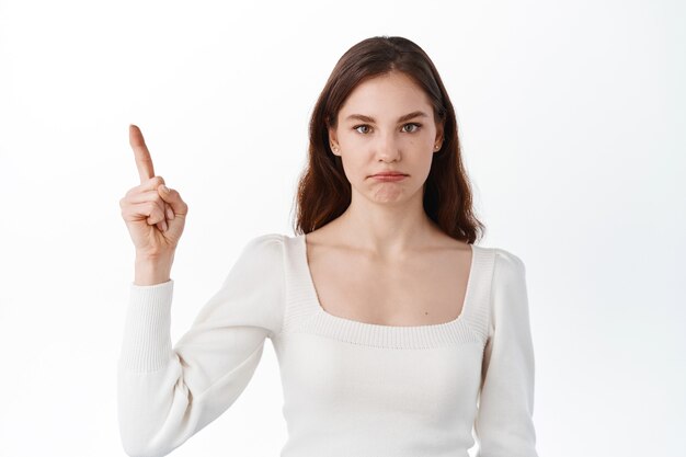 Disappointed and sad girl pointing at unfair bad thing, points up at logo on top and pouting upset, standing displeased against white wall