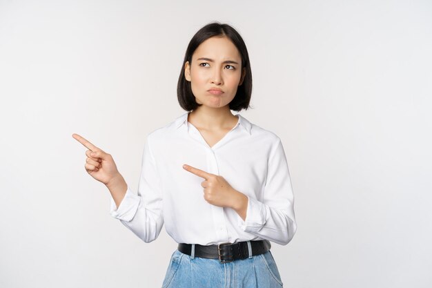 Disappointed sad young asian woman pointing and looking left with upset sulking face expression standing over white background