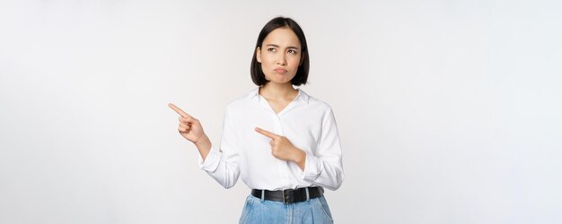 Disappointed sad young asian woman pointing and looking left with upset sulking face expression standing over white background