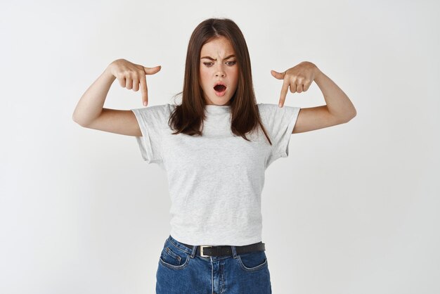 Disappointed and shocked young woman staring pointing fingers down at bad banner express dislike and frustration standing over white background