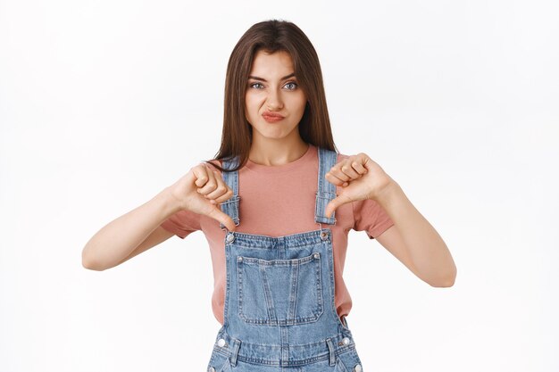 Disappointed skeptical arrogant attractive woman in overalls, t-shirt, smirking unsatisfied, showing thumbs-down and frowning in disagreement, express dislike judge something awful