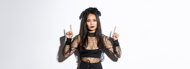 Disappointed and skeptical asian female in black dress and wreath looking displeased, pointing fingers up at something bad, have negative opinion, standing over white background