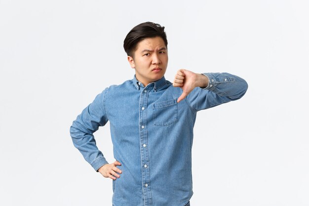 Disappointed skeptical asian man looking judgemental and unamused, standing white background shaking head and showing thumbs-down in disapproval, dislike and dont recommend something