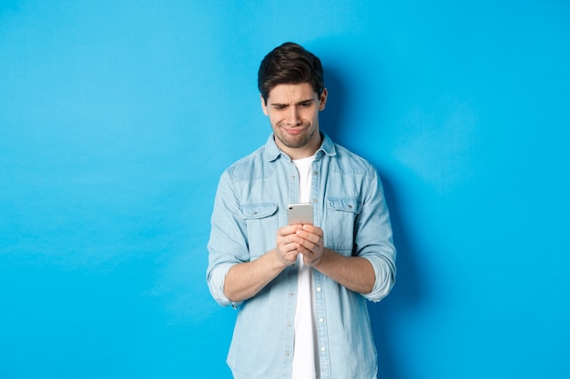 Disappointed and skeptical guy looking at phone, standing over blue background