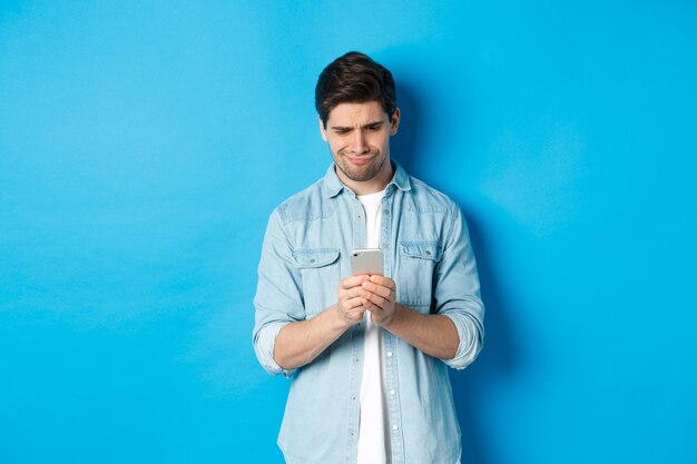 Disappointed and skeptical guy looking at phone, standing over blue background