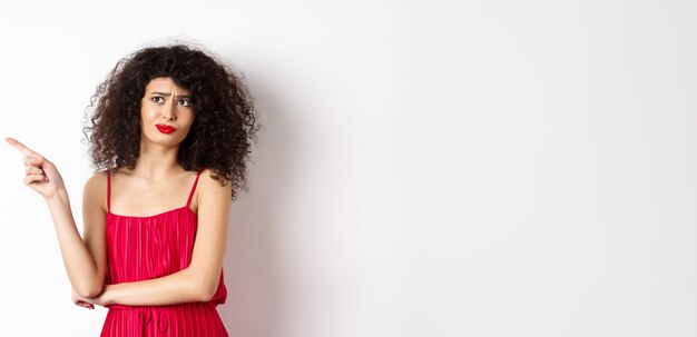 Free photo disappointed and skeptical young woman with curly hair wearing red dress grimacing and pointing fing