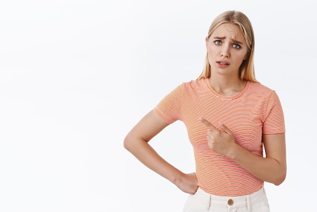 Foto gratuita delusa e leggermente sconvolta ragazza bionda carina cupa che si lamenta riluttante a fare il dovere quotidiano mentre indica a sinistra chiedendo a qualcuno di coprirle le sopracciglia curve e solcate dispiaciuto sfondo bianco