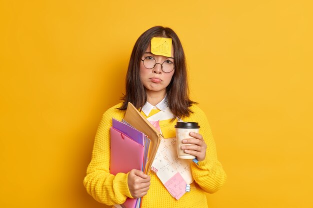 실망한 피곤한 여성 졸업생이 시험 세션을 준비하고 마감일이 스티커 메모로 둘러싸여 있고 종이가 일회용 커피를 보유하고 있습니다.
