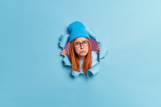 Разочарованная расстроенная молодая женщина поджимает губы с оскорбительным выражением лица, грустно смотрит с длинными рыжими волосами, носит шляпу и высовывает голову из разорванной голубой бумажной стены.