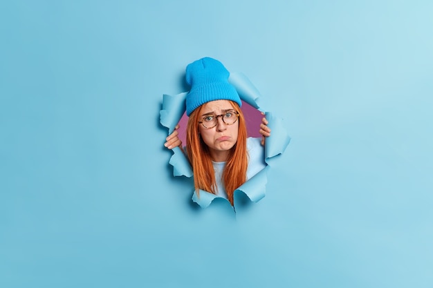 Disappointed upset young woman purses lips with offensive expression looks sadly has long red hair wears hat and transparent glasses sticks head out of ripped hole blue paper wall.