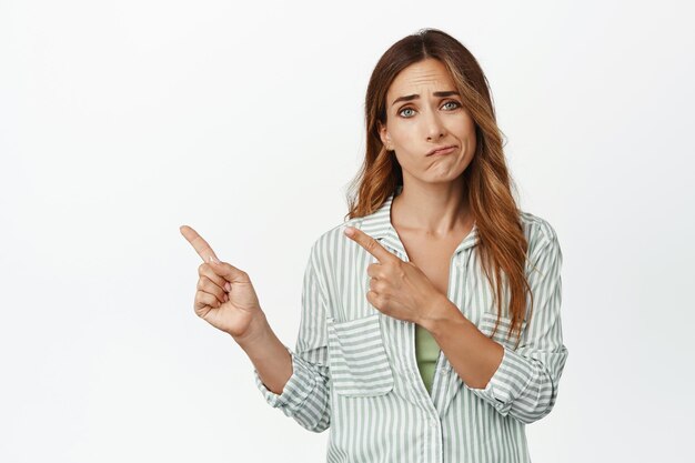 Disappointed woman frowning upset, feel pity sad, pointing fingers left with sadness, standing skeptical reluctant against white background