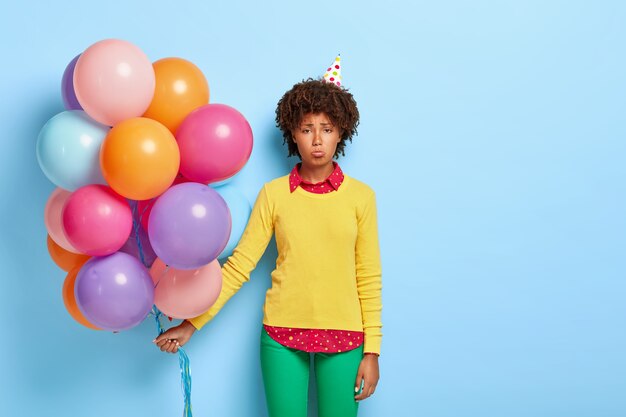 Disappointed woman holds multicolored balloons while posing in a yellow sweater