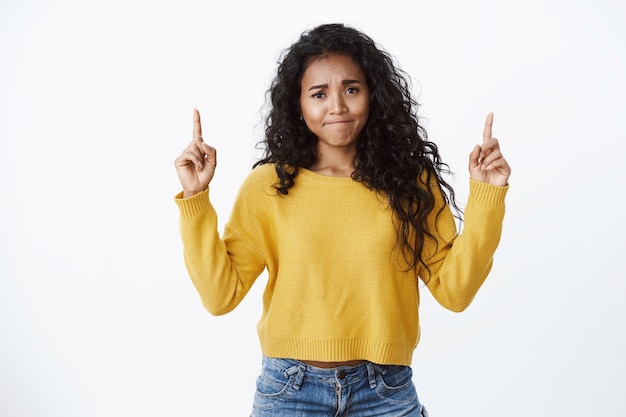 Giovane ragazza afroamericana delusa che increspa le labbra aggrottando la fronte e puntando verso l'alto insoddisfatta, dando un feedback negativo, sembrando scettica e indifferente, in piedi maglione giallo muro bianco