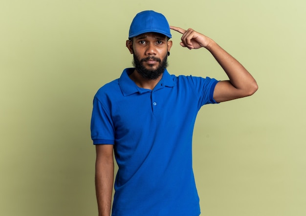 Disappointed young afro-american delivery man pointing at his cap isolated on olive green wall with copy space