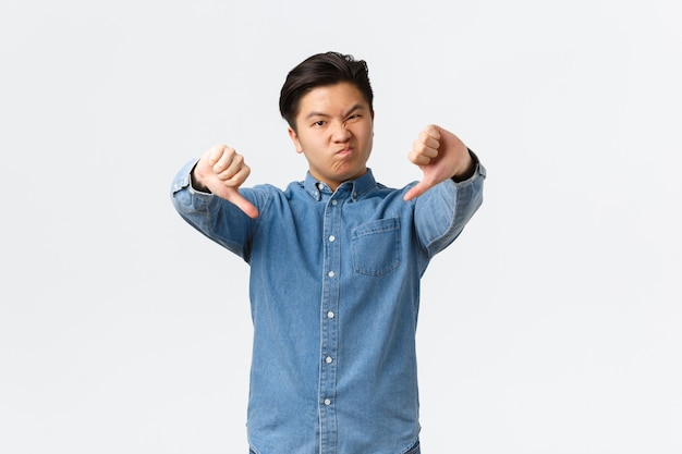 Disappointed young asian man complaining on bad quality, showing thumbs-down and grimacing displeased, left unamused, dislike and disagree, standing white background.
