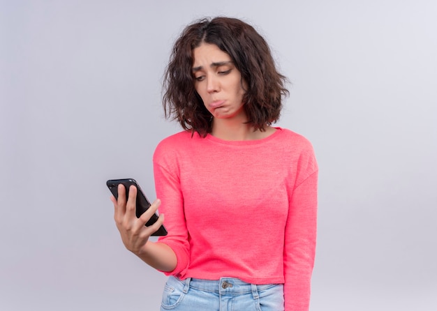 Disappointed young beautiful woman holding mobile phone and looking at it on isolated white wall