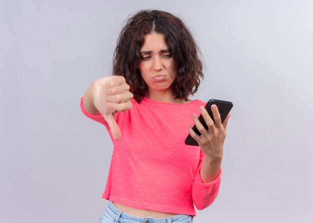 Disappointed young beautiful woman holding mobile phone and showing thumb down on isolated white wall