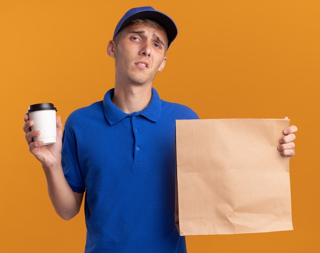 Disappointed young blonde delivery boy holds paper package and cup isolated on orange wall with copy space