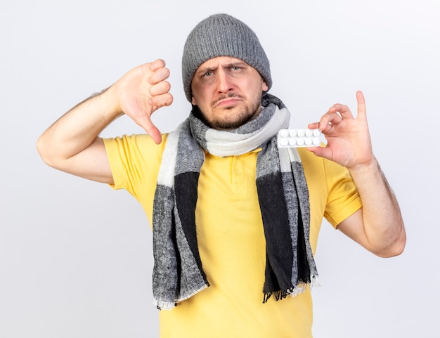 Foto gratuita il giovane uomo slavo malato biondo deluso che indossa il cappello e la sciarpa di inverno i pollici giù e tiene il pacchetto delle pillole mediche isolate sulla parete bianca con lo spazio della copia