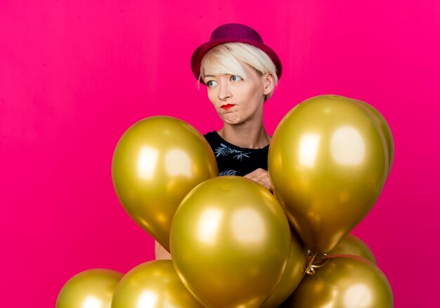 Disappointed young blonde party girl wearing party hat standing behind balloons looking at side isolated on crimson background