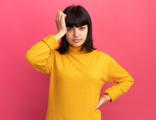 Disappointed young brunette caucasian girl puts hand on head isolated on pink wall with copy space