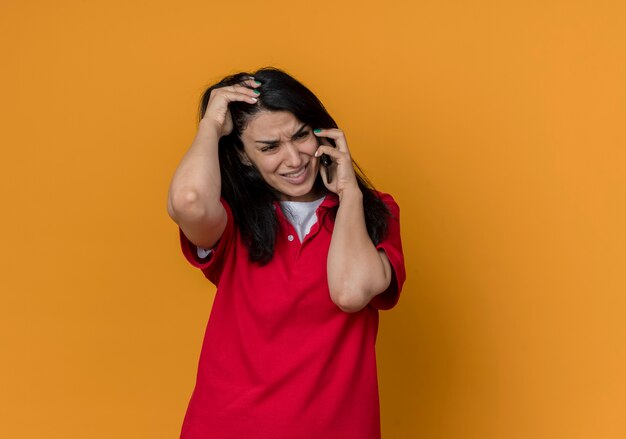 Disappointed young brunette caucasian girl wearing red shirt talks on phone and holds head isolated on orange wall