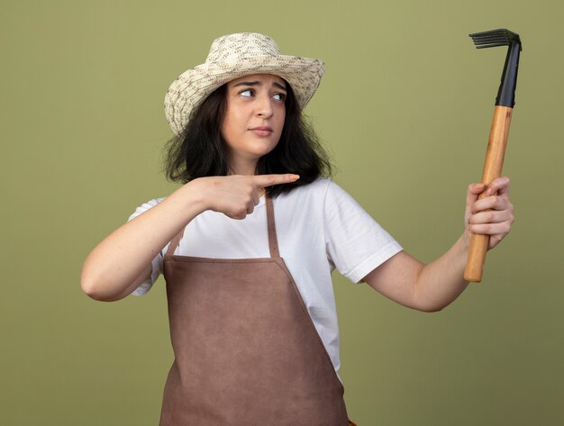 Disappointed young brunette female gardener in uniform wearing gardening hat holds and points at rake isolated on olive green wall