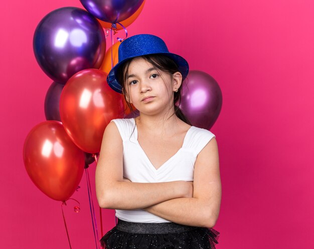 disappointed young caucasian girl with blue party hat standing with crossed arms in front of helium balloons isolated on pink wall with copy space