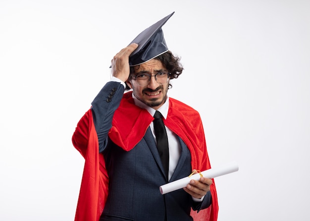 赤いマントを着たスーツを着て、卒業証書を持っている卒業式の帽子に手を置いて、眼鏡をかけたがっかりした若い白人のスーパーヒーローの男