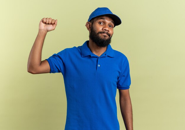 disappointed young delivery man standing with raised fist isolated on olive green wall with copy space