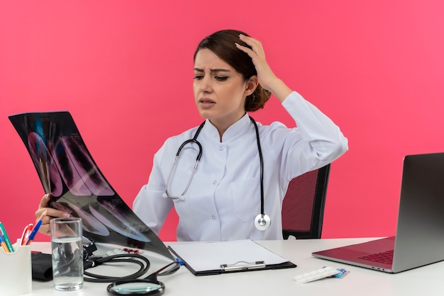 Disappointed young female doctor wearing medical robe and stethoscope sitting at desk with medical tools and laptop holding and looking at x-ray shot touching head 