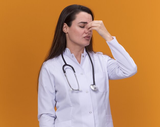 Disappointed young female doctor wearing medical robe with stethoscope puts hand on nose isolated on orange wall with copy space