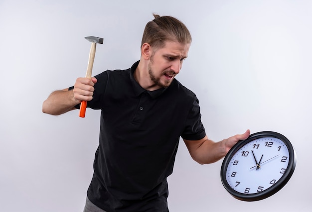 Disappointed young handsome man in black polo shirt holding wall clock and swinging a hammer going to break the clock standing over white background