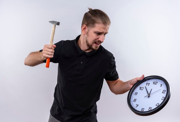 Разочарованный молодой красавец в черной рубашке поло держит настенные часы и размахивает молотком, собираясь сломать часы, стоящие на белом фоне