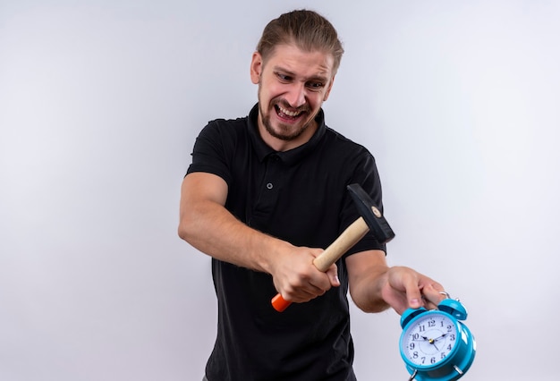 Disappointed young handsome man in black polo shirt with a hammer going to break alarm clock in his hand with aggressive expression on face standing over white background