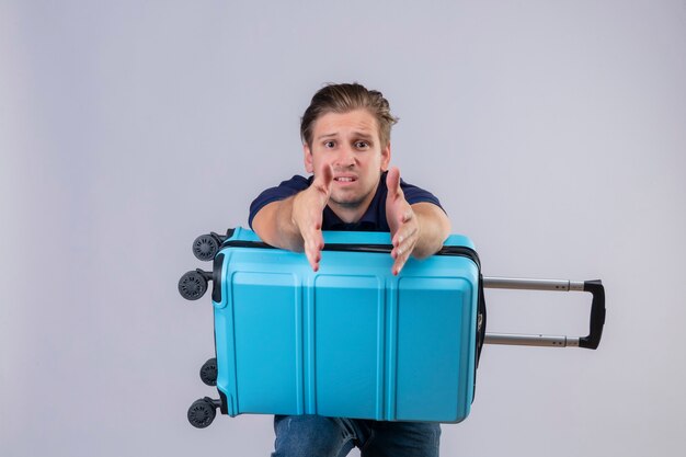 Disappointed young handsome traveler guy standing with suitcase stretching hands out  asking for help 