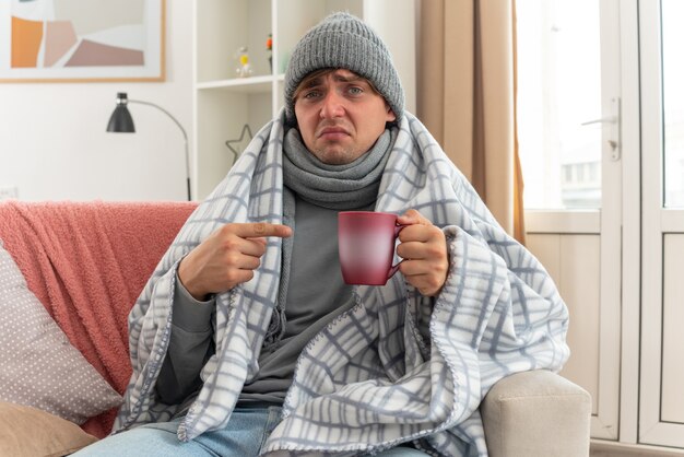 disappointed young ill man with scarf around neck wearing winter hat wrapped in plaid holding and pointing at cup sitting on couch at living room
