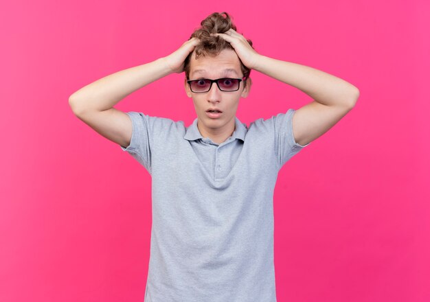 Disappointed young man in black glasses wearing grey polo shirt touching his head being confused standing over pink wall