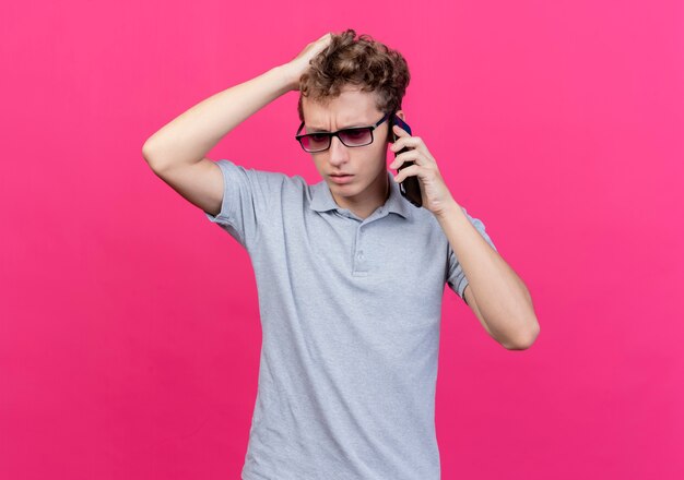 Disappointed young man in black glasses wearing grey polo shirt touching his head being confused while talking on mobile phone standing over pink wall