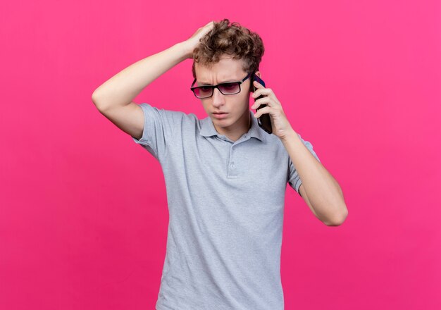 ピンクの壁の上に立っている携帯電話で話しているときに混乱している彼の頭に触れている灰色のポロシャツを着て黒い眼鏡をかけた失望した若い男