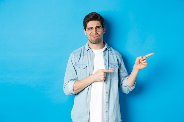 Disappointed young man in glasses pointing fingers right at copy space, showing promo banner and smirking dissatisfied, judging bad product, standing over blue background.