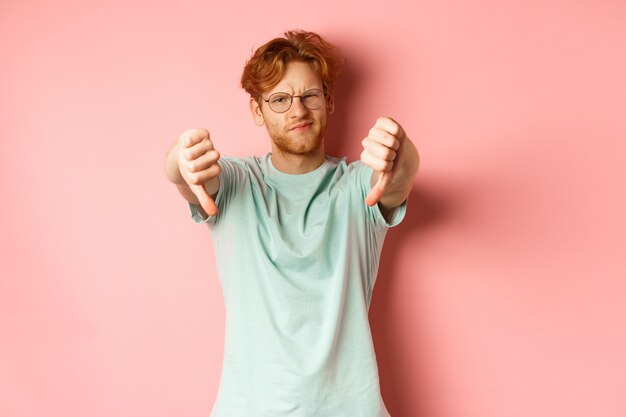 Disappointed young man in glasses with messy red haircut showing thumbs down and grimacing unsatisfi...