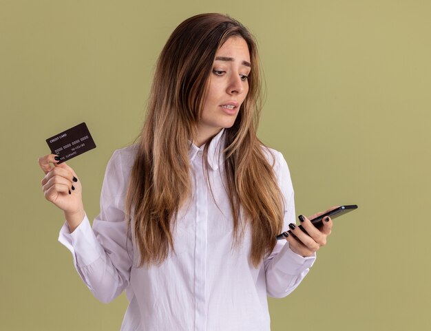 失望した若いかなり白人の女の子はクレジットカードを持って電話を見る