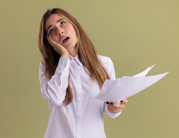 Disappointed young pretty caucasian girl puts hand on face and holds blank paper sheets looking up