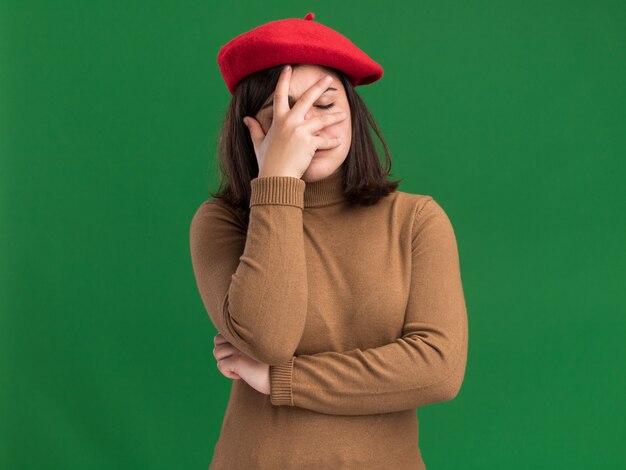 Disappointed young pretty caucasian girl with beret hat puts hand on face on green