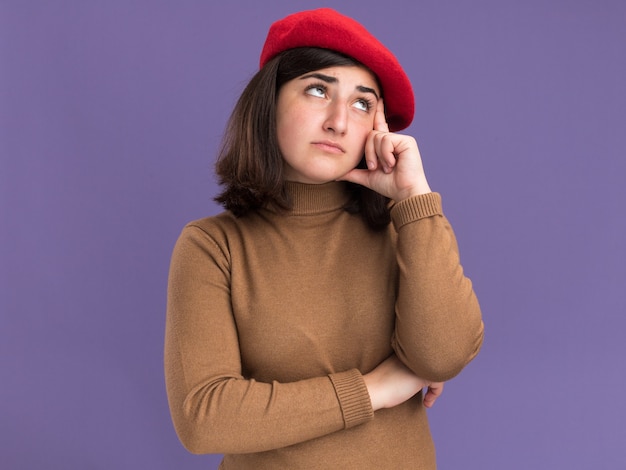 Free photo disappointed young pretty caucasian girl with beret hat puts hand on temple and looks up on purple