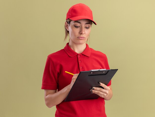 Disappointed young pretty delivery woman in uniform writes on clipboard with pen isolated on olive green wall