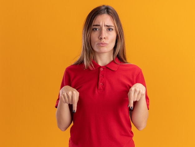 Разочарованная молодая красивая женщина показывает палец вниз двумя руками, изолированными на оранжевой стене